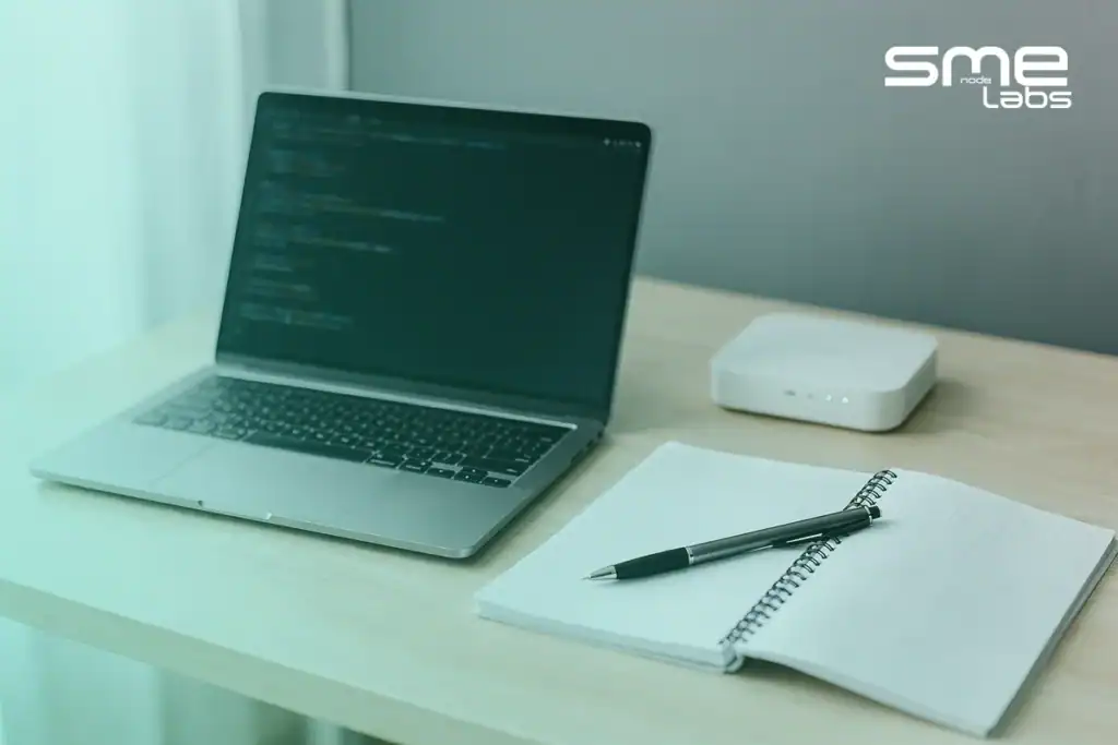 Laptop with a terminal, a spiral workbook and a router on a desk, representing the MikroTik MTCNA Workbook labs