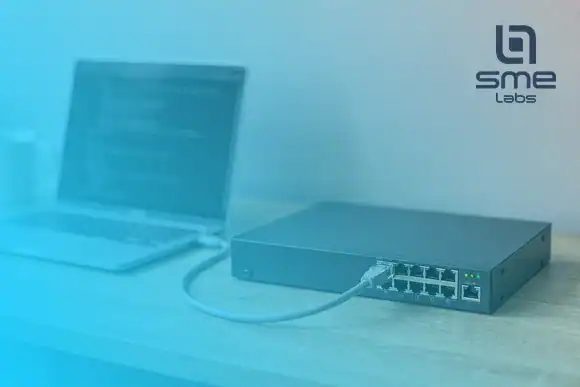 An open laptop on a wooden bench joined by one grey cable to a small dark network switch with green status lights, under a light blue fade from the bottom-left corner.
