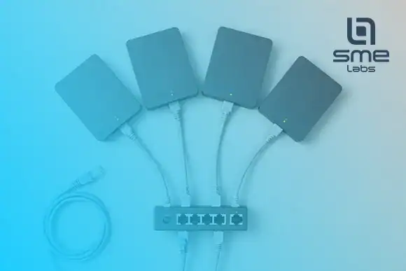 Four charcoal desktop routers on a light grey desk, each joined by one grey cable to a small switch, with a light blue console cable coiled nearby, under a light blue fade from the bottom-left corner.