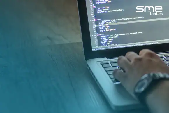 A hand typing code on a laptop on a wooden desk, with SMEnode Labs cyan fading in from the bottom-left corner.