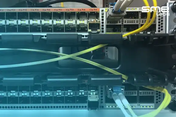 Two rack-mounted fibre switches, one above the other, linked by fibre patch cables, with SMEnode Labs cyan fading in from the bottom-left corner.