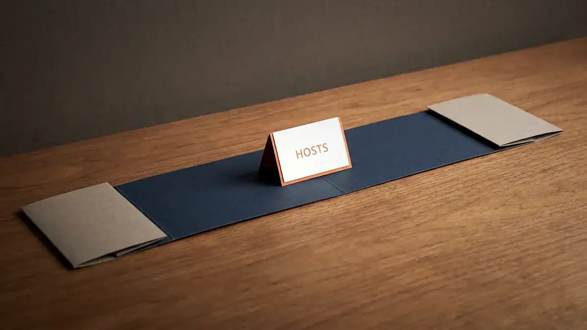 A paper strip on a wooden table with short grey segments at both ends and a long navy middle segment holding a card that reads HOSTS.