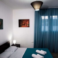 Aegean View Villa – Υπνοδωμάτιο με διπλό κρεβάτι, γαλάζιες κουρτίνες και πίνακες τοίχου