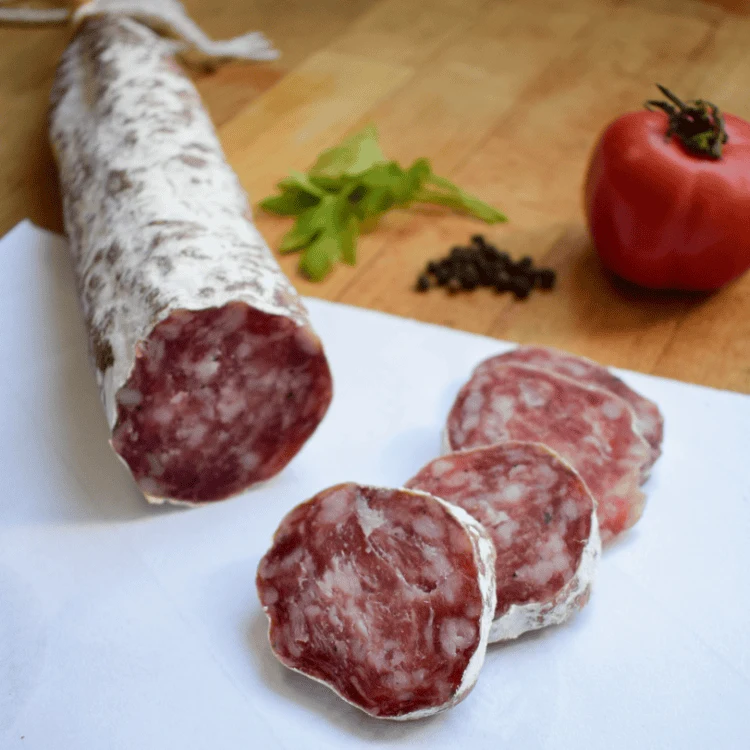 Sliced salami on a white surface with a whole salami log, tomato, and herbs in the background on a wooden board.