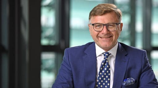 Mann in Anzug und Brille vor moderner Bürofensterfront.