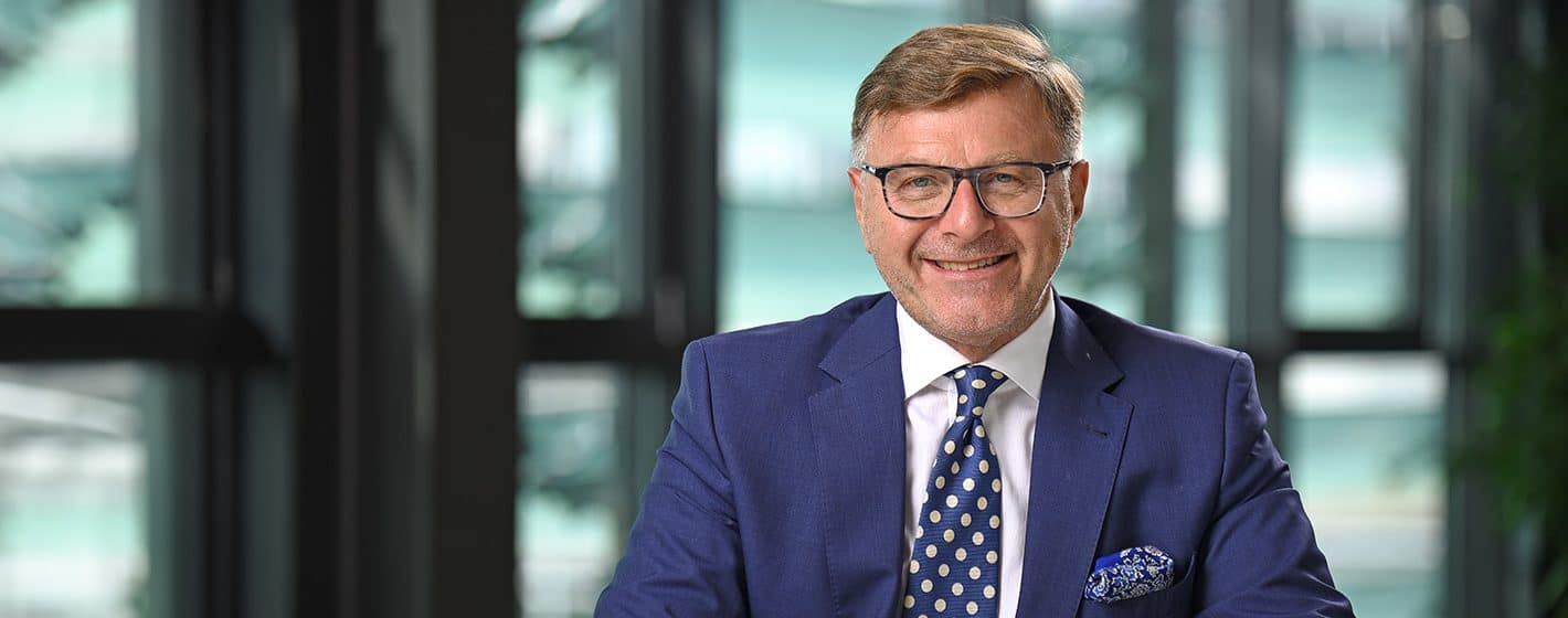 Mann in Anzug und Brille vor moderner Bürofensterfront.