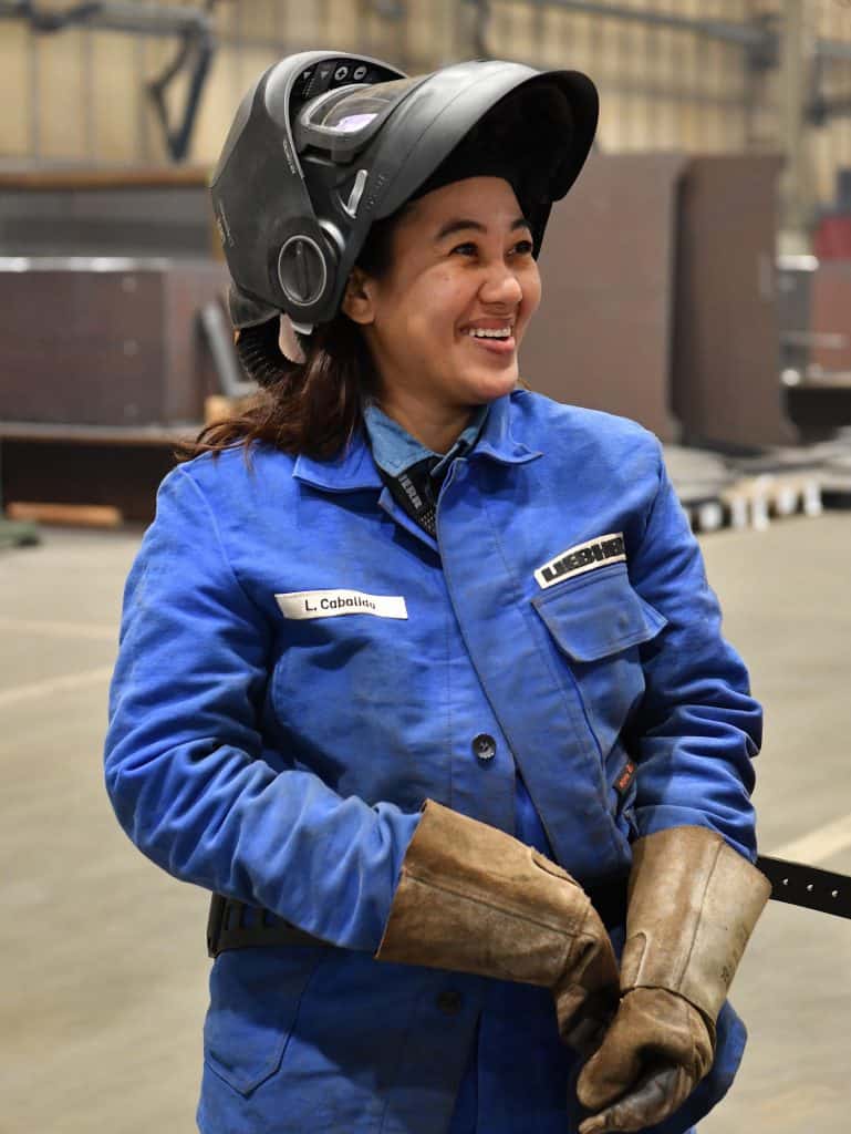 Frau in blauer Arbeitsjacke mit Schutzhelm und Handschuhen im Industrieumfeld.
