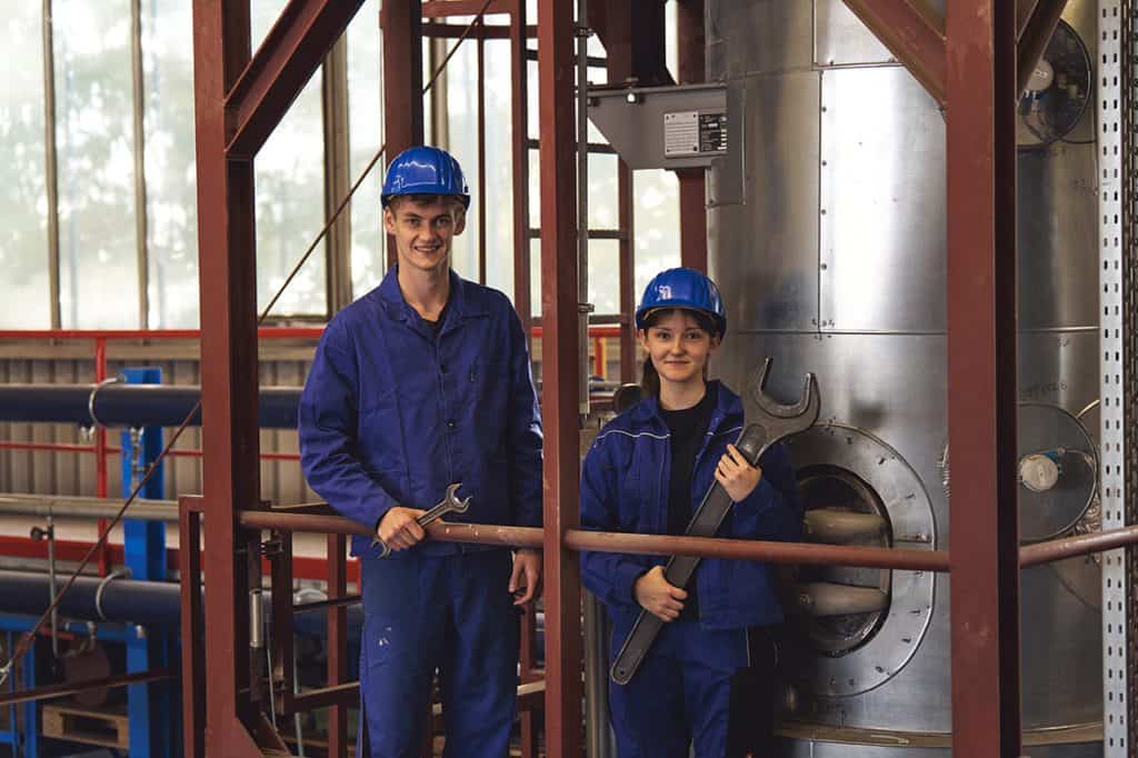 Zwei junge Handwerker in blauen Arbeitsanzügen und Helmen bei der Arbeit.