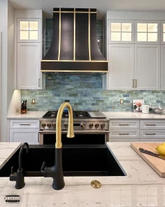 Kitchen with light countertops, blue tile backsplash, and black range hood with gold accents