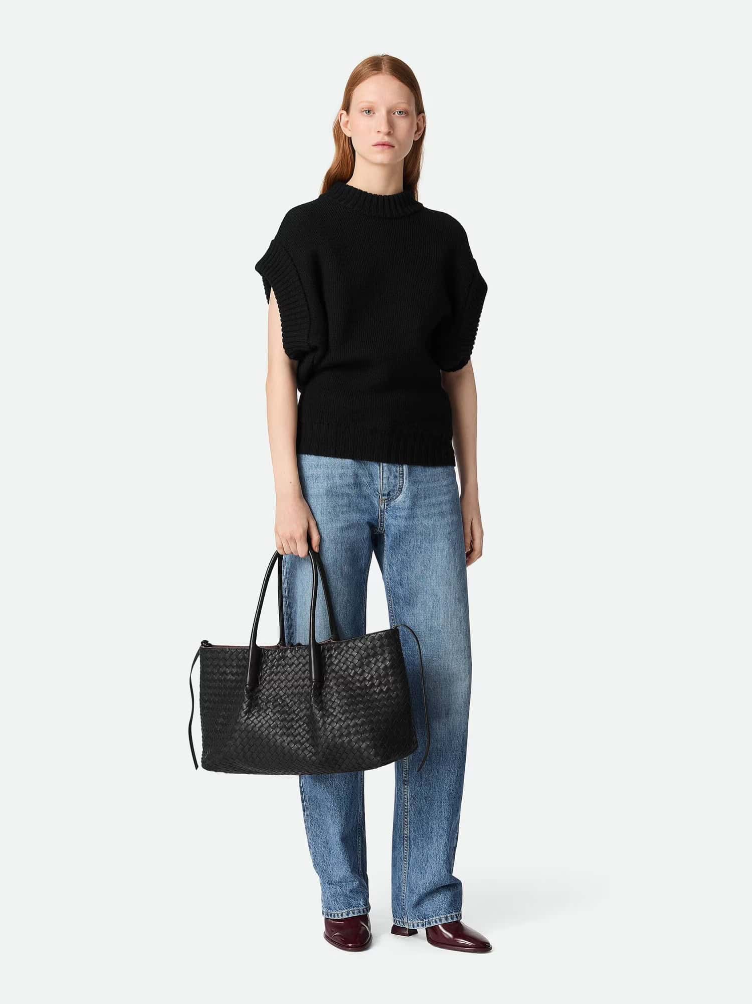 Mulher usando casaco preto de malha com jeans e bota escura, segurando uma bolsa de couro preta.