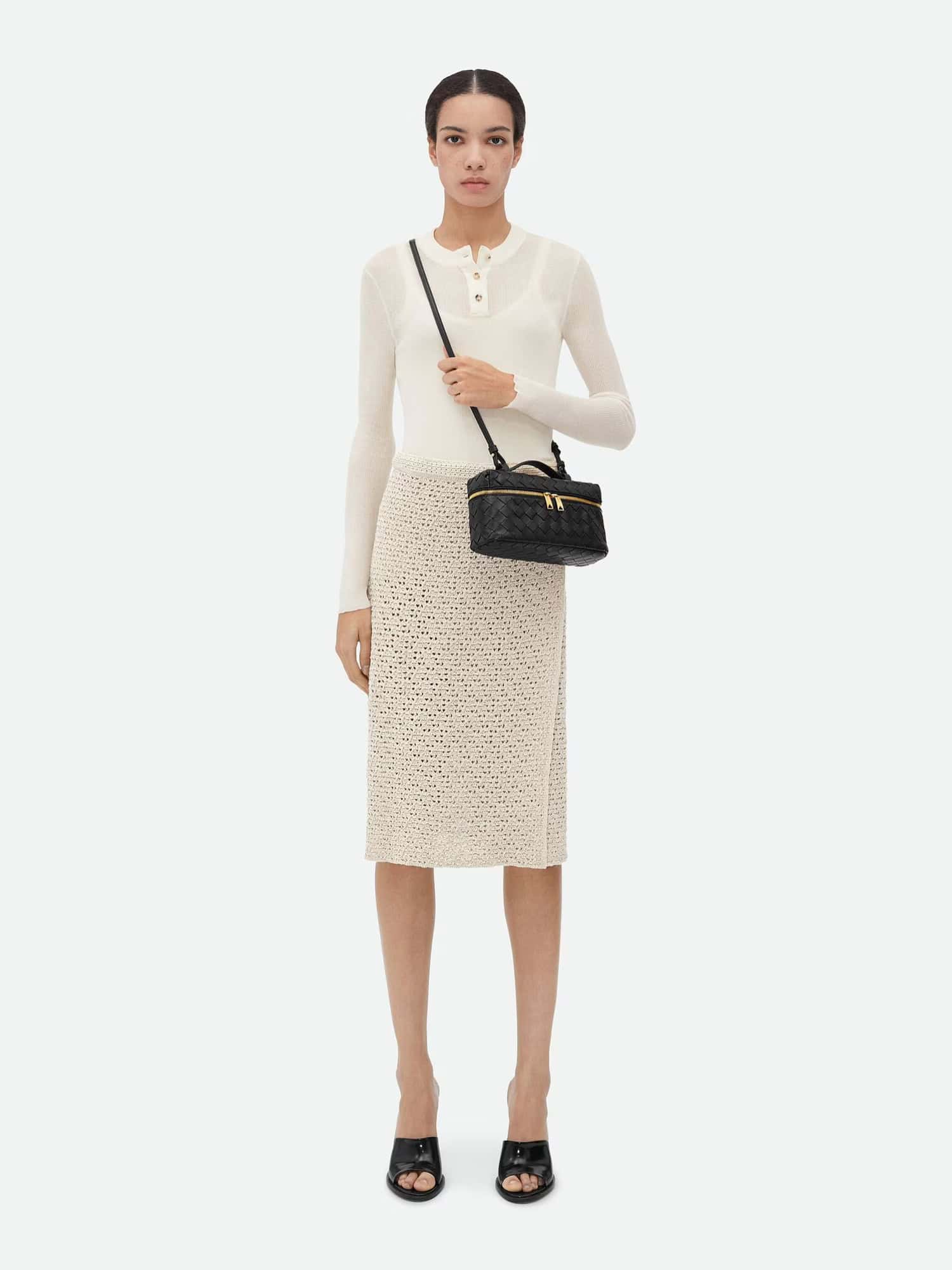Elegante mulher com saias de crochê e top de manga comprida, carregando uma bolsa preta em um fundo branco.