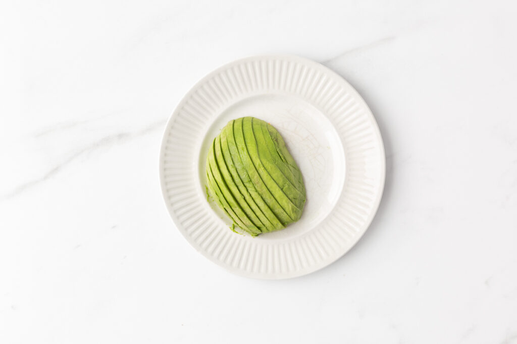 Process shot #1: Half an avocado with the flesh scooped out and slice lengthwise into thin slices. Placed on a small plate.
