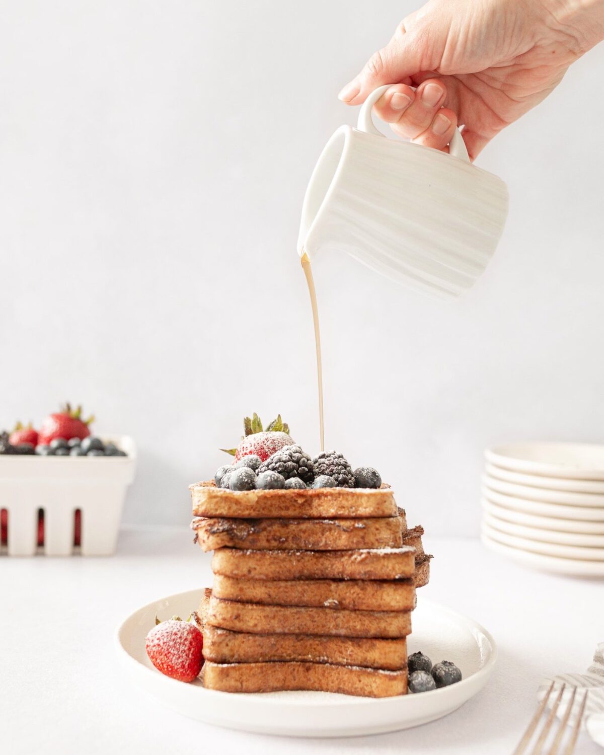 Classic French Toast stacked high on a plate and topped with fresh berries. Maple syrup is being drizzled on top.