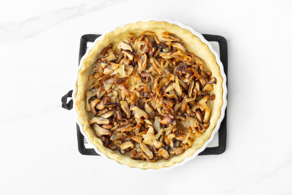 Process shot #3: Caramelized onions and mushrooms spread out in an even layer in the bottom of a prepared quiche crust.