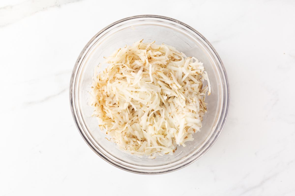 Process shot #2: Shredded potato with the liquid squeezed out. Potatoes transferred to a glass mixing bowl.