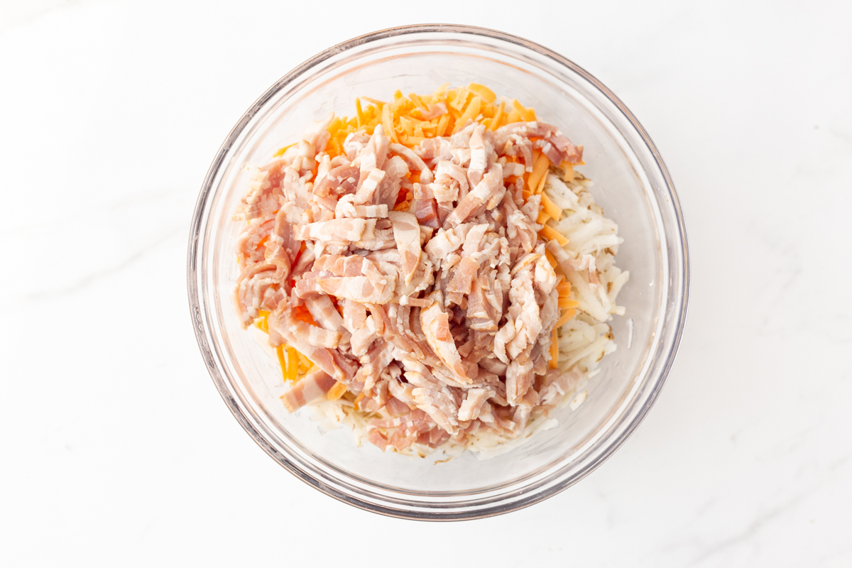 Process shot #3: A glass mixing bowl with shredded potatoes, shredded cheddar cheese and diced bacon in it.