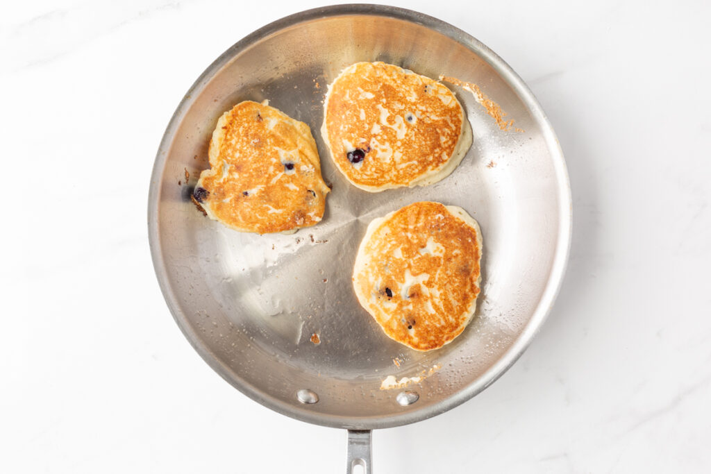 Process shot #4: 3 cooked blueberry pancakes in a stainless steel skillet.