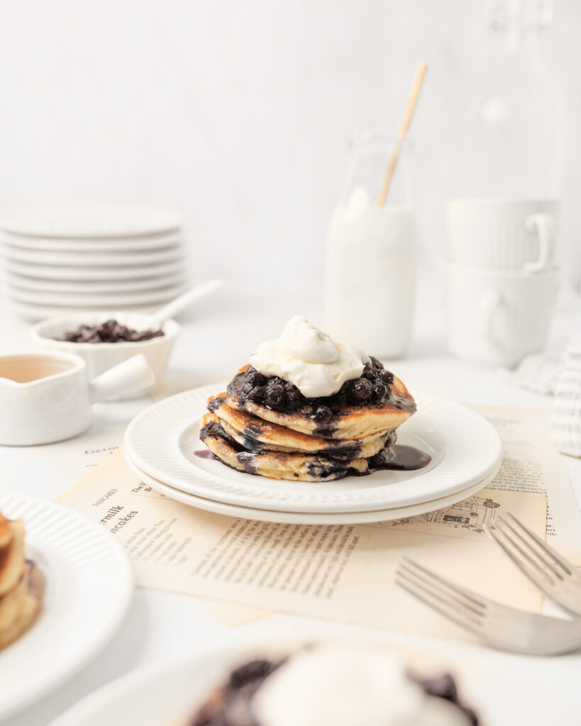 3 gluten free blueberry pancakes stacked on a small plate and topped with blueberry sauce and whipped cream.