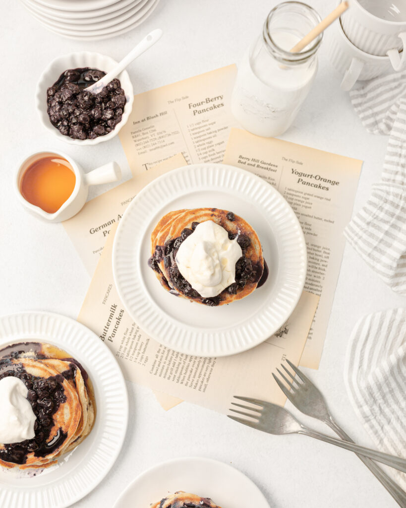Gluten Free Blueberry Pancakes served on a small plate with blueberry sauce and whipped cream. There is syrup on the side.