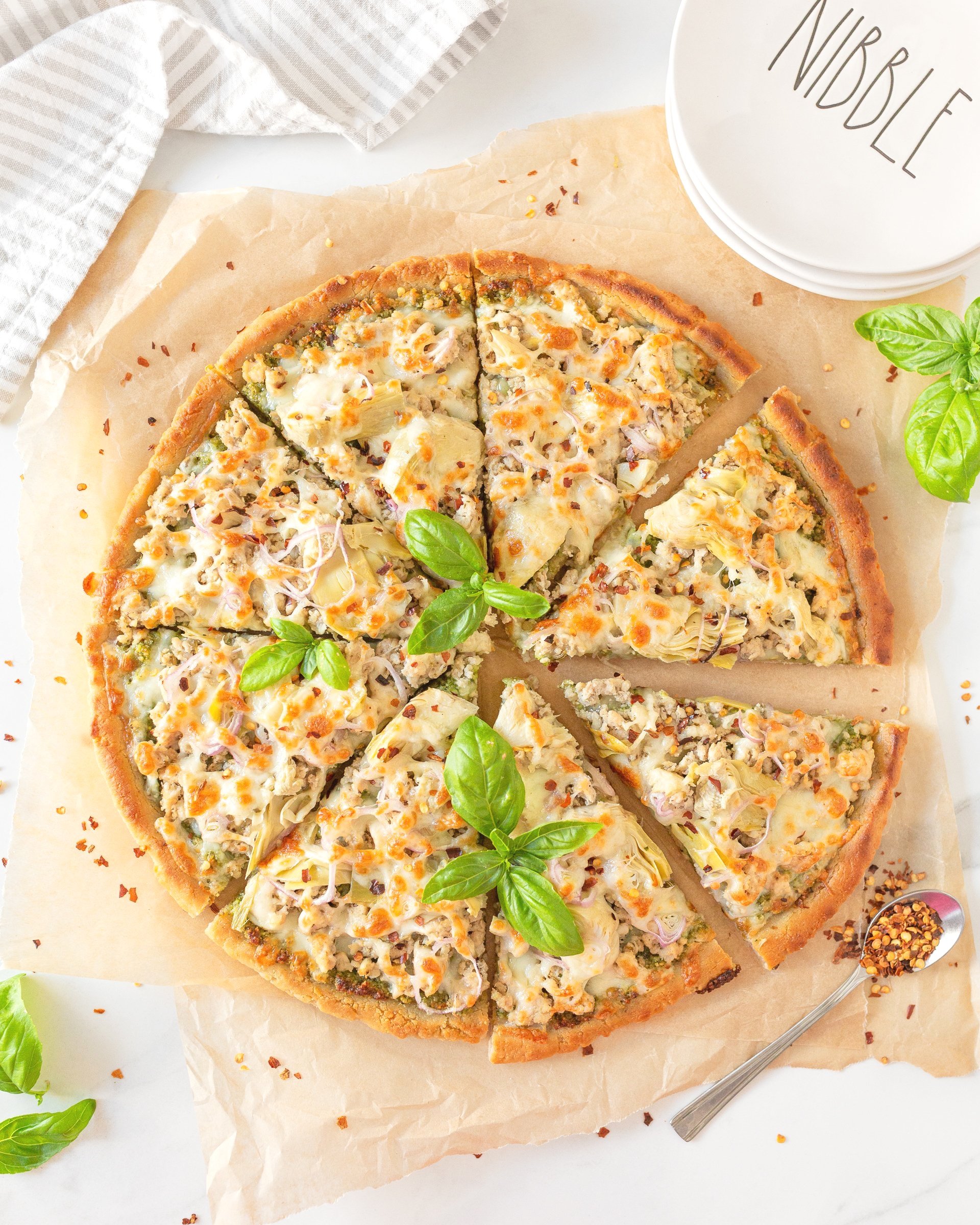 Ground Chicken Pesto Pizza with Artichoke Hearts cut on parchment paper and garnished with fresh basil.