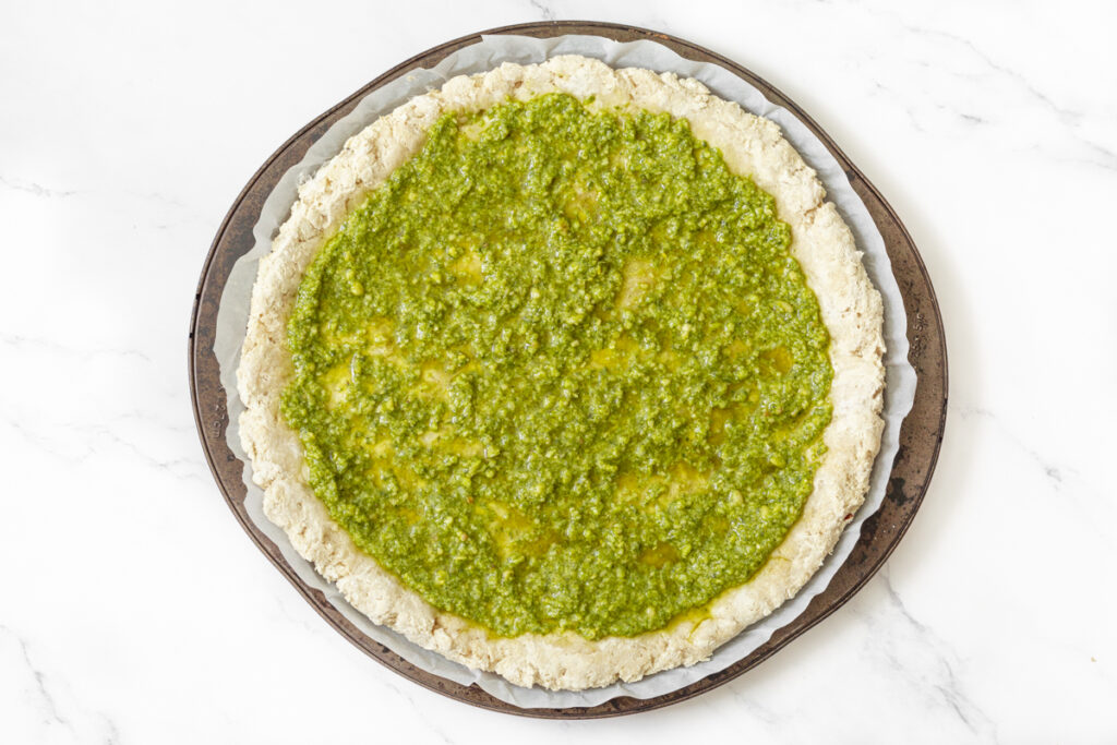Process shot #2: A pre-baked gluten free pizza crust on a pizza pan with a generous layer on basil pesto spread on top.
