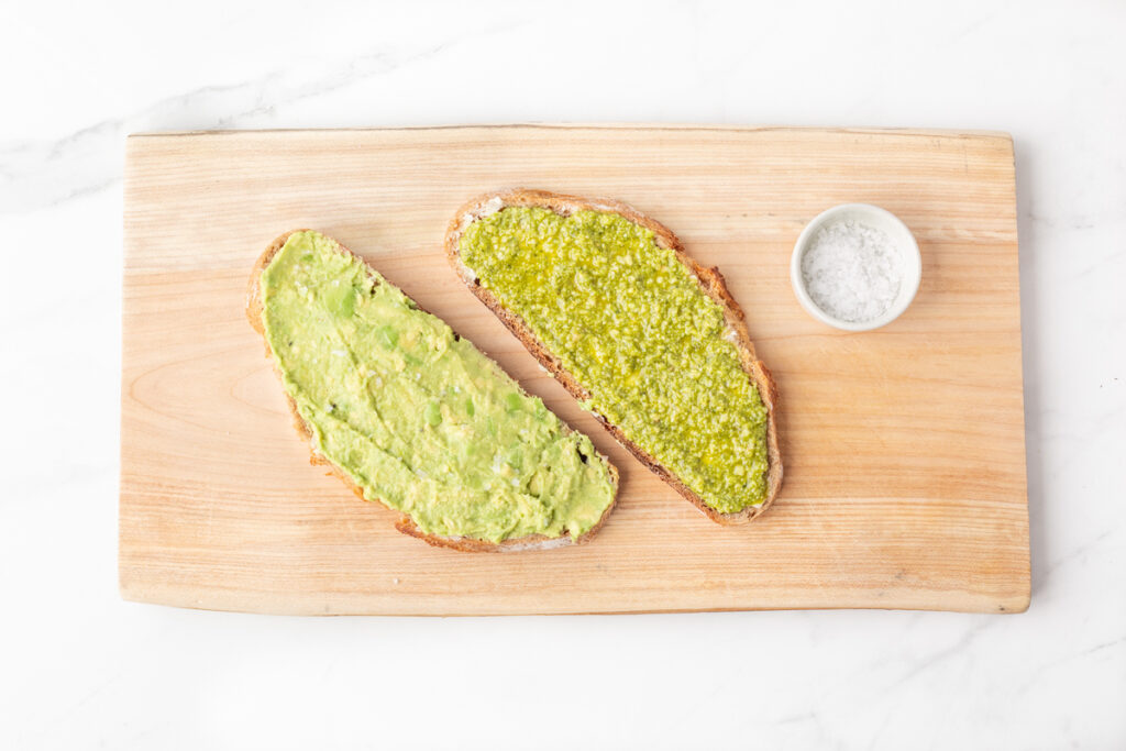 Process shot #3: Two slices of sourdough bread, one with mashed avocado spread on top and the other with basil pesto spread on top.