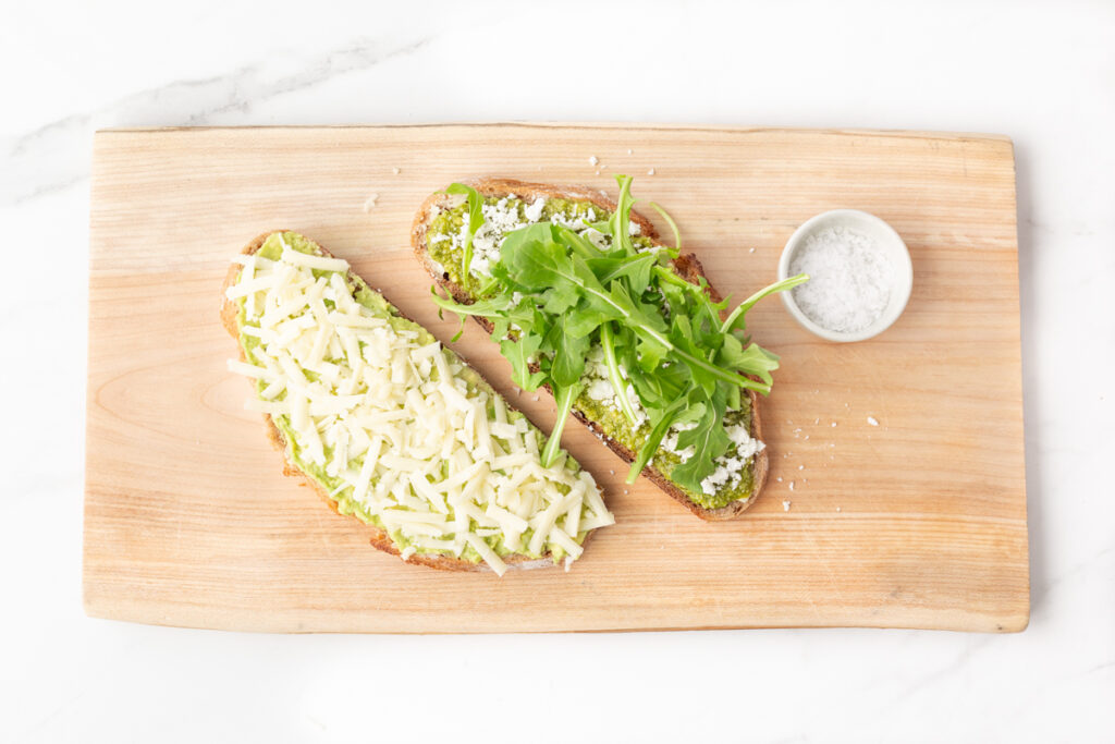 Process shot #4: Two slices of sourdough bread: one with pesto, feta cheese and arugula and the other with mashed avocado and shredded mozzarella cheese.