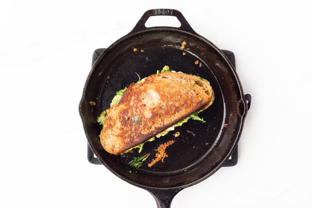 Process shot #5: Grilled pesto, avocado and feta sandwich crispy and golden brown in a cast iron pan.
