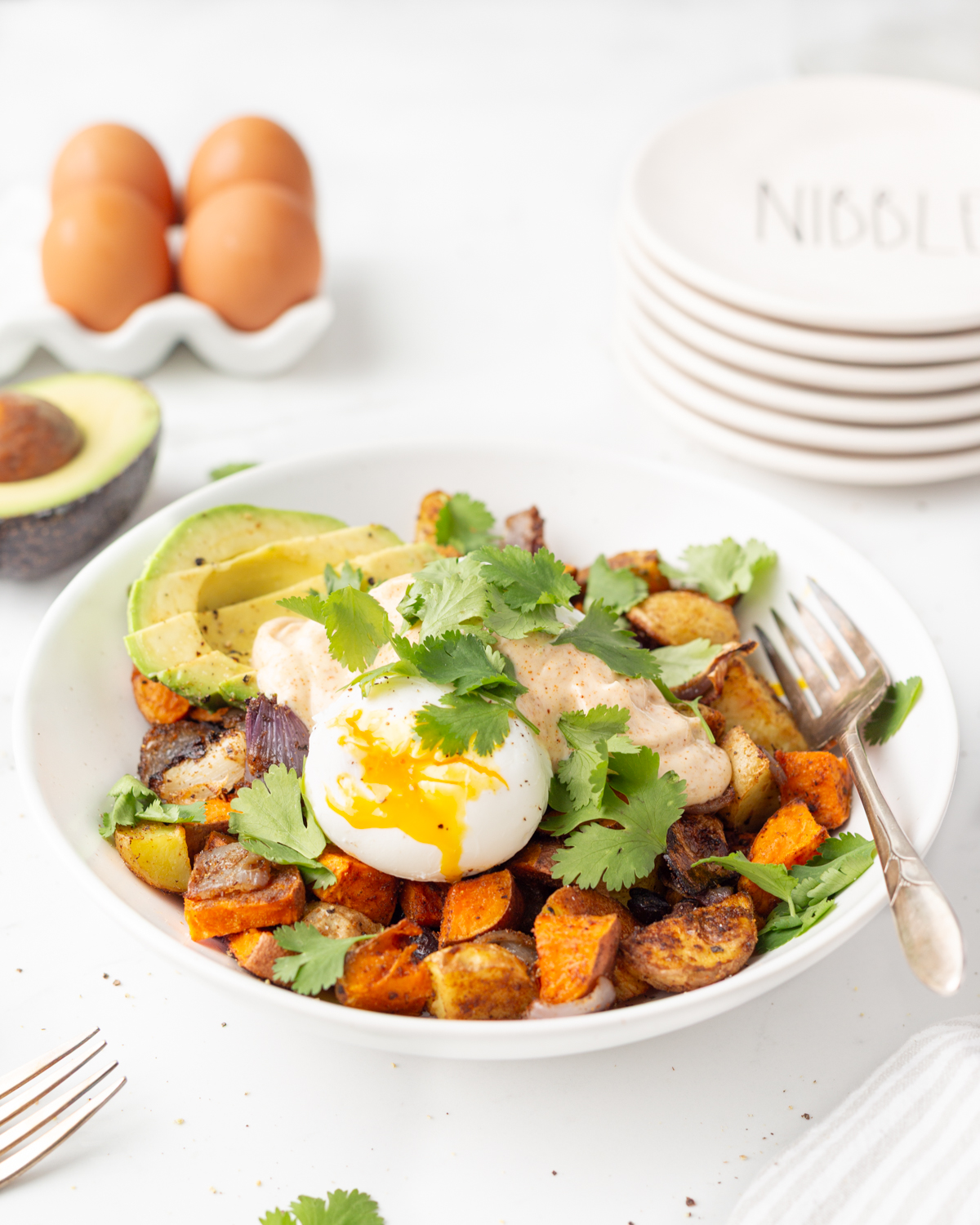 Process shot #3: Southwest Breakfast Bowl plated including roasted potato mixture topped with sliced avocado, poached eggs, chipotle mayo and fresh cilantro.