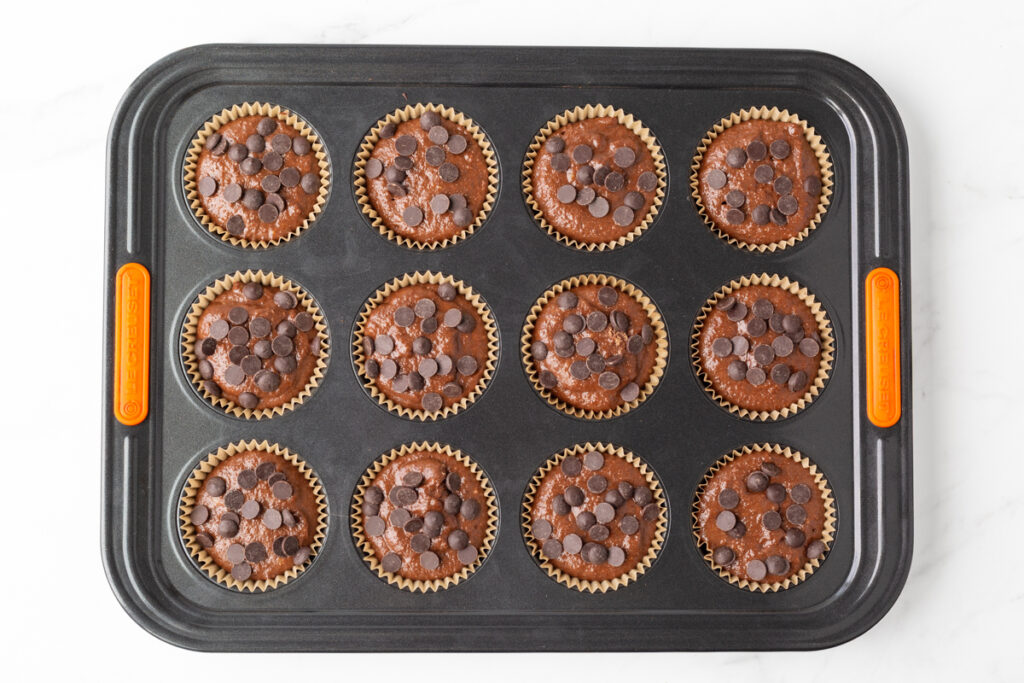 Process shot #4: Chocolate Banana Muffin batter evenly scooped into 12 paper liners in a muffin tin and topped with chocolate chips.