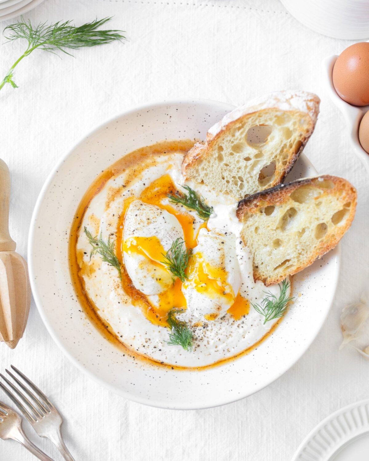 Creamy Turkish Eggs in a bowl topped with chilli butter and fresh dill. Crusty sourdough bread slices on the side.