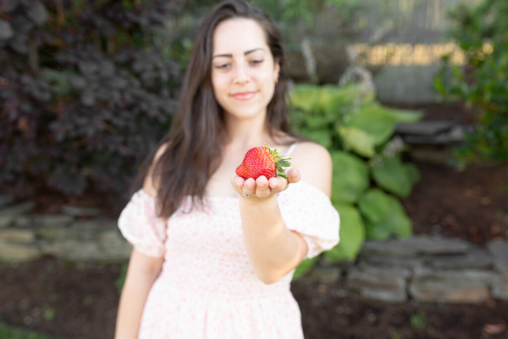 Courtney holding a giant strawberry up to the camera. She is outdoors.