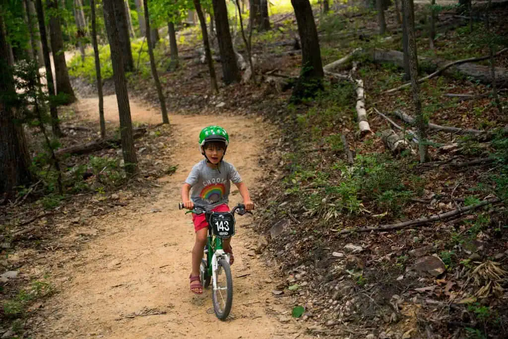 Child in a green helmet riding a small bike along a dirt trail through the woods