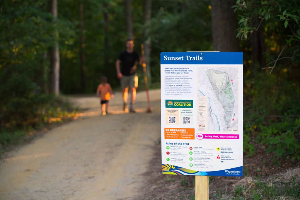 Families walking past a Sunset Park trail map sign at the edge of the woods