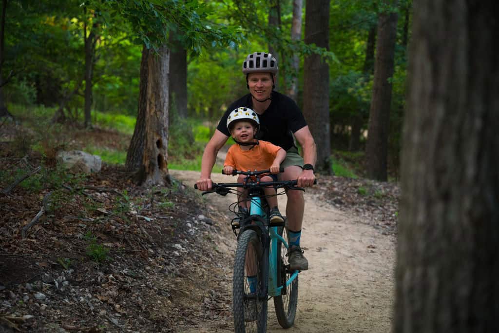 Parent riding a mountain bike with a young child seated up front on a wooded Sunset Park trail