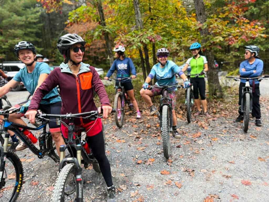 A group of smiling riders on mountain bikes gathered at a gravel trailhead under fall foliage, ready for a Coalition group ride