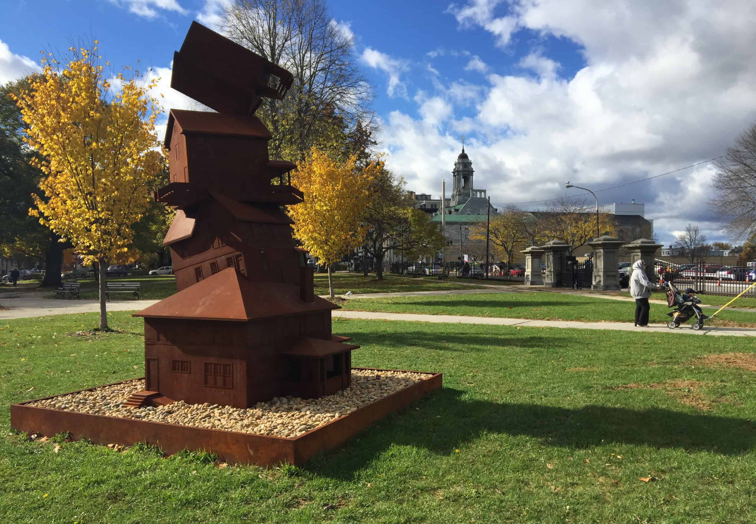 Contemporary rust-colored metal sculpture in a park with trees and city skyline.