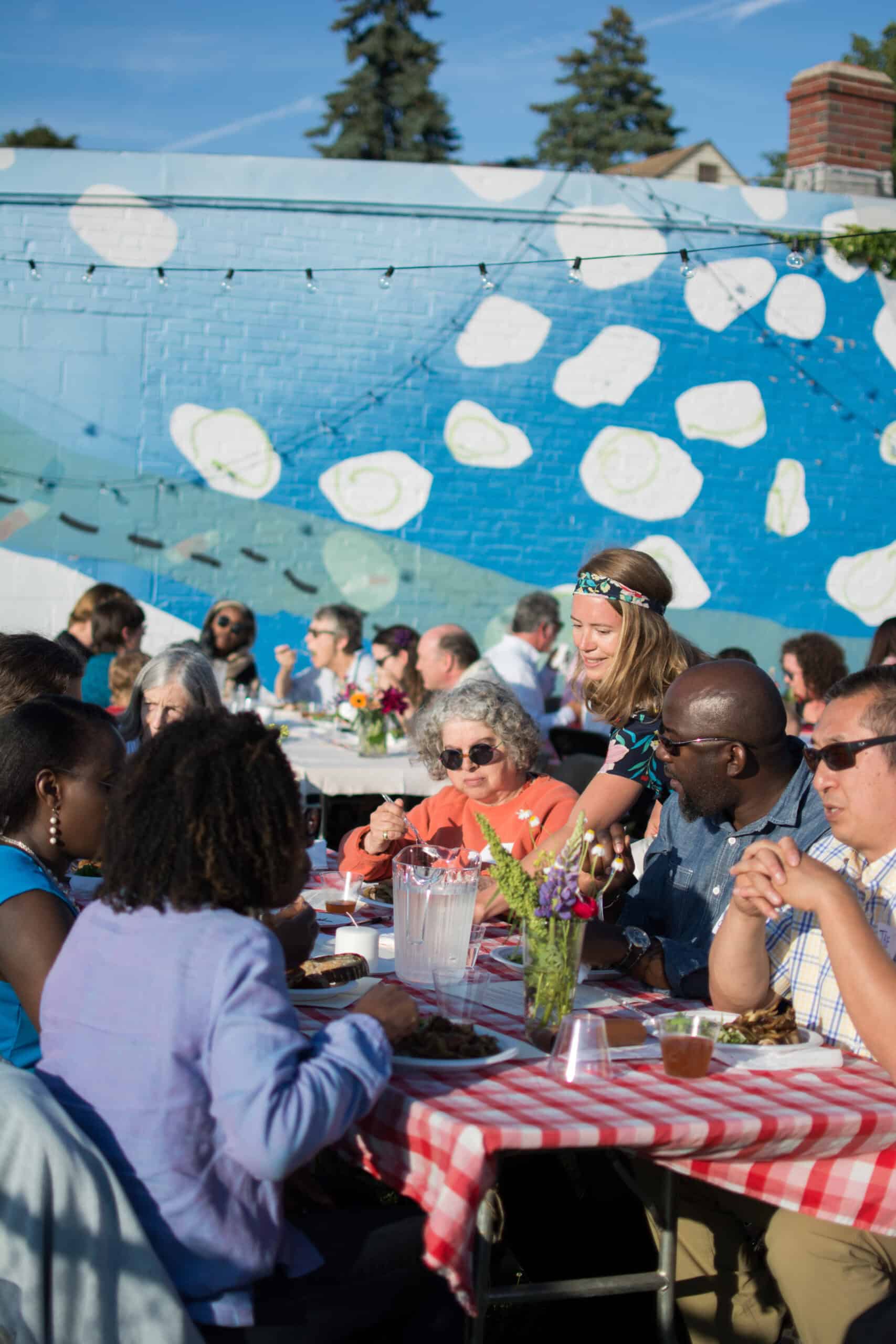 Outdoor community event with diverse attendees dining at a table in front of a colorful mural.