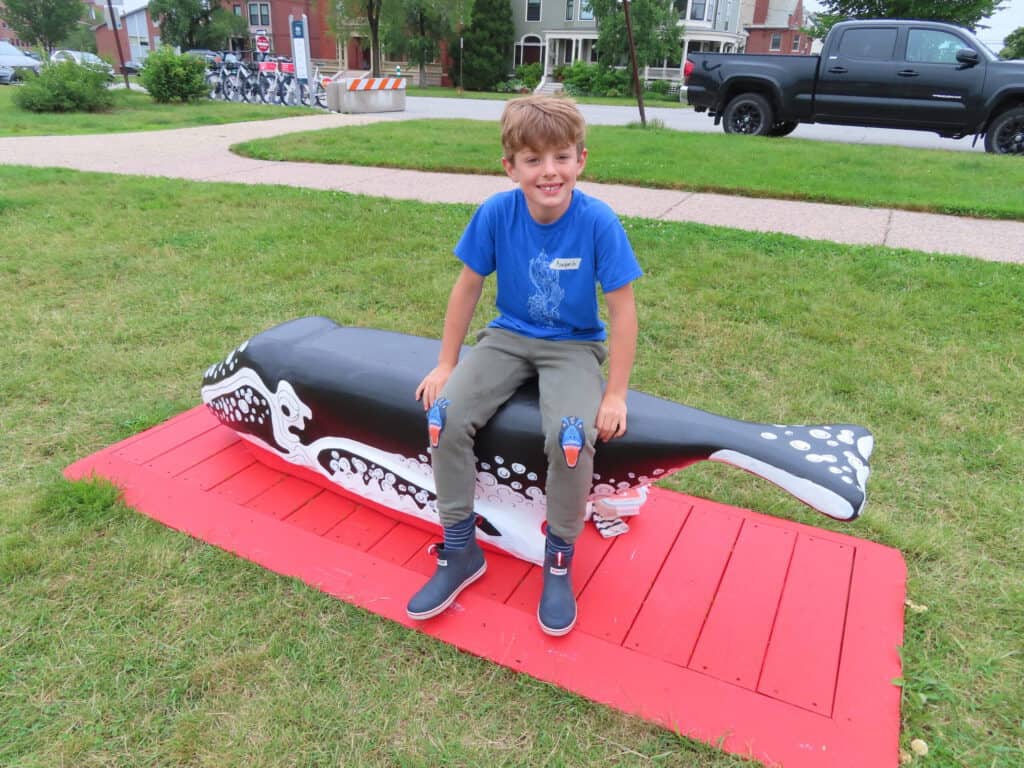 Child sitting on a large painted whale sculpture in a park.