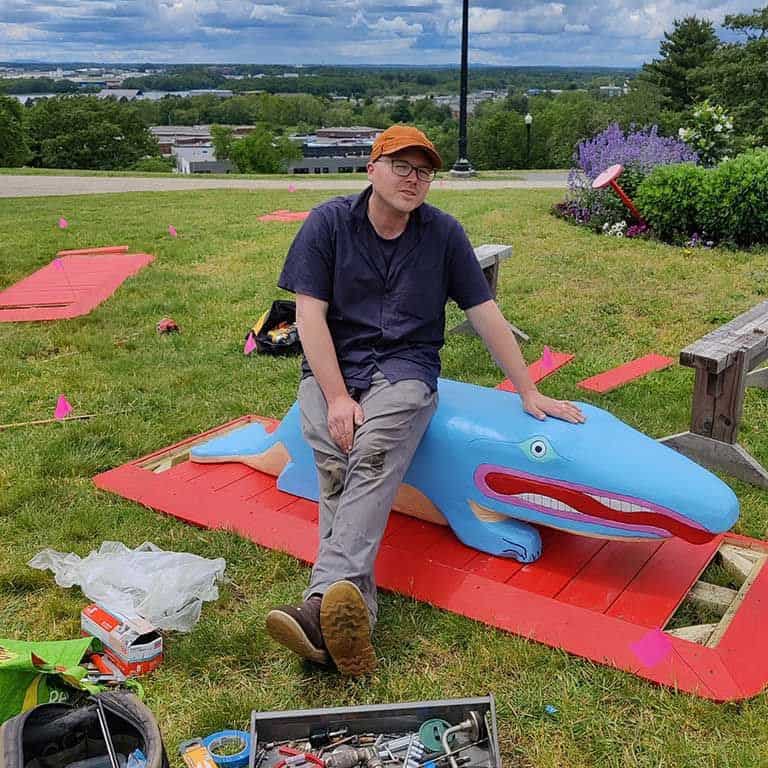 Person sitting on a colorful crocodile sculpture in a park during summer 2023.