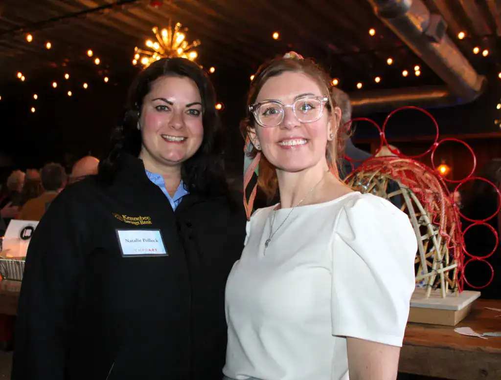 Two women smiling at an art event with vibrant lighting and creative installations in the background.