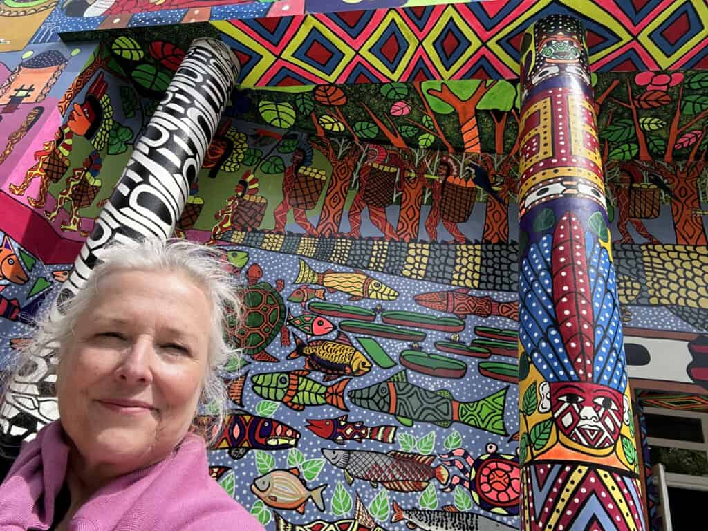 An image of Sue Lambe in front of a colorful, intricate mural celebrating community and diversity.