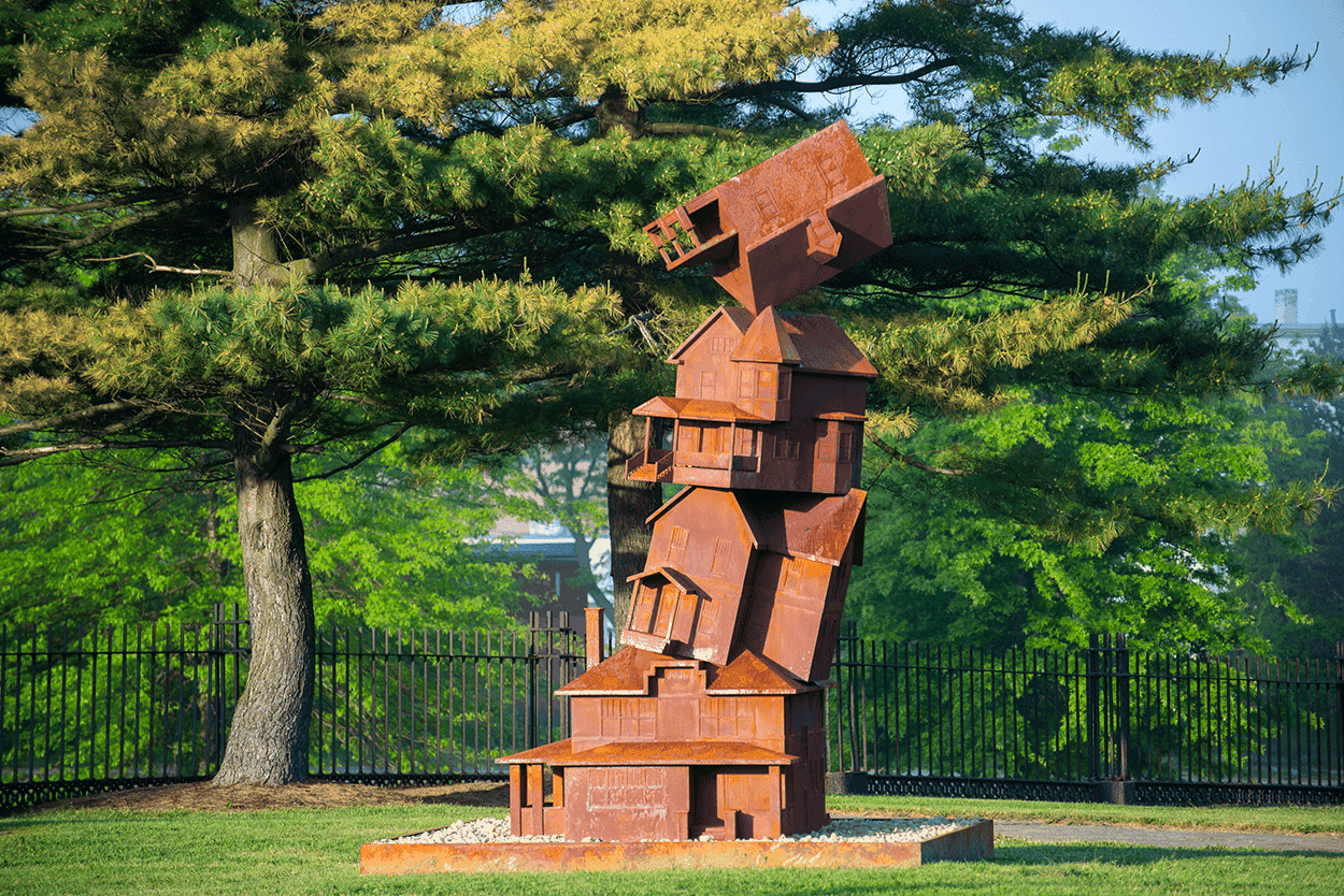 Rust-colored abstract metal sculpture in a lush green park with trees and a black fence in the backg.