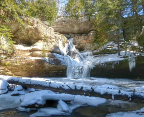 Snow-covered waterfall in a forested area, winter landscape, cold weather scene, frozen water, scenic outdoor nature.