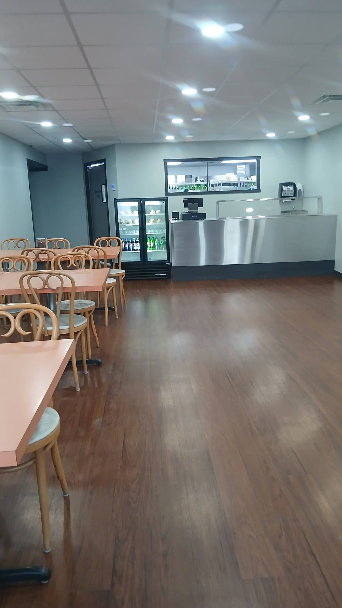 Empty cafe dining area with wooden tables and chairs, modern decor, and refrigerated drinks behind the counter.