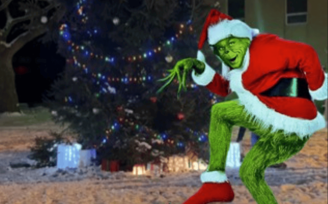 Festive Grinch dressed as Santa poses near decorated Christmas tree with gifts, holiday spirits.
