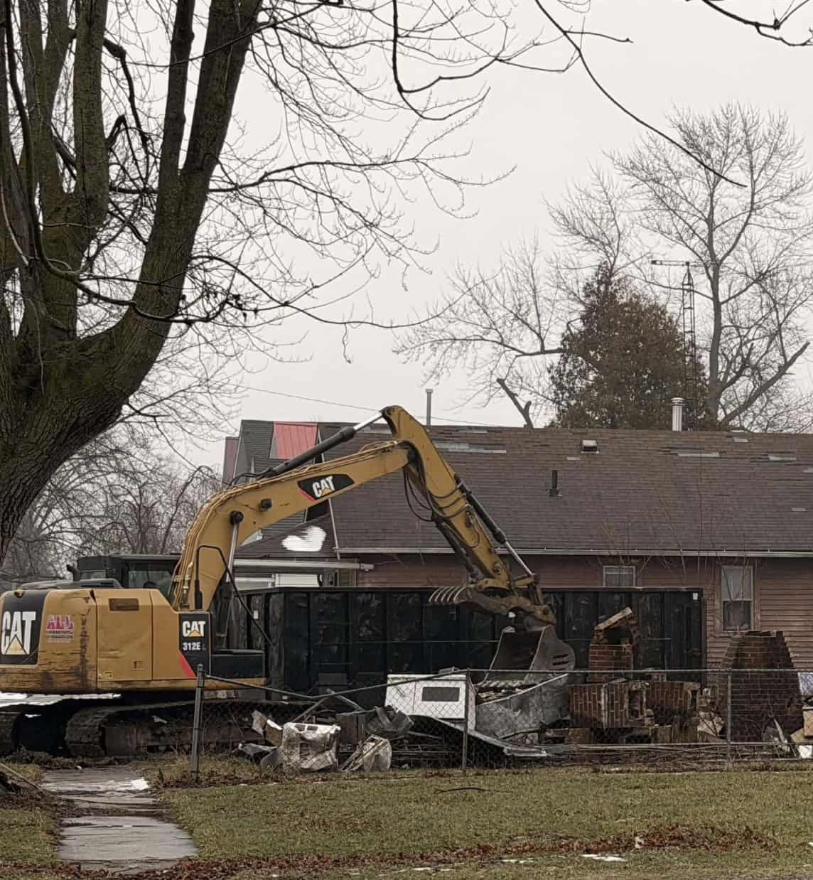 Heavy construction equipment demolishing a house in Fostoria, Ohio, on a cloudy day.