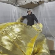 Golden sculpture artist working on a large gold-colored art piece at an outdoor event.