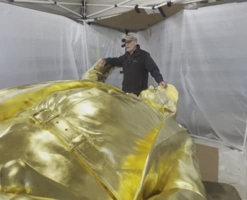 Golden sculpture artist working on a large gold-colored art piece at an outdoor event.