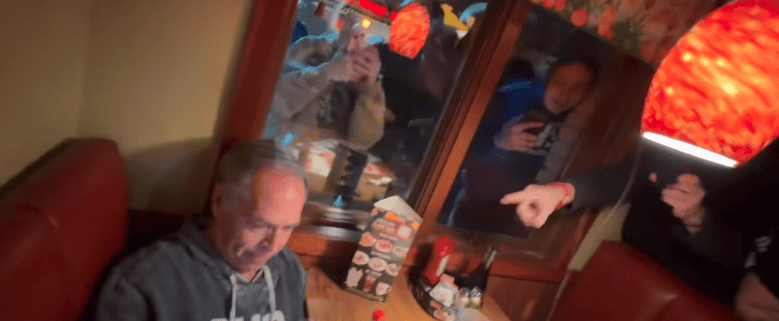 Man using phone in restaurant with people pointing and taking photos.